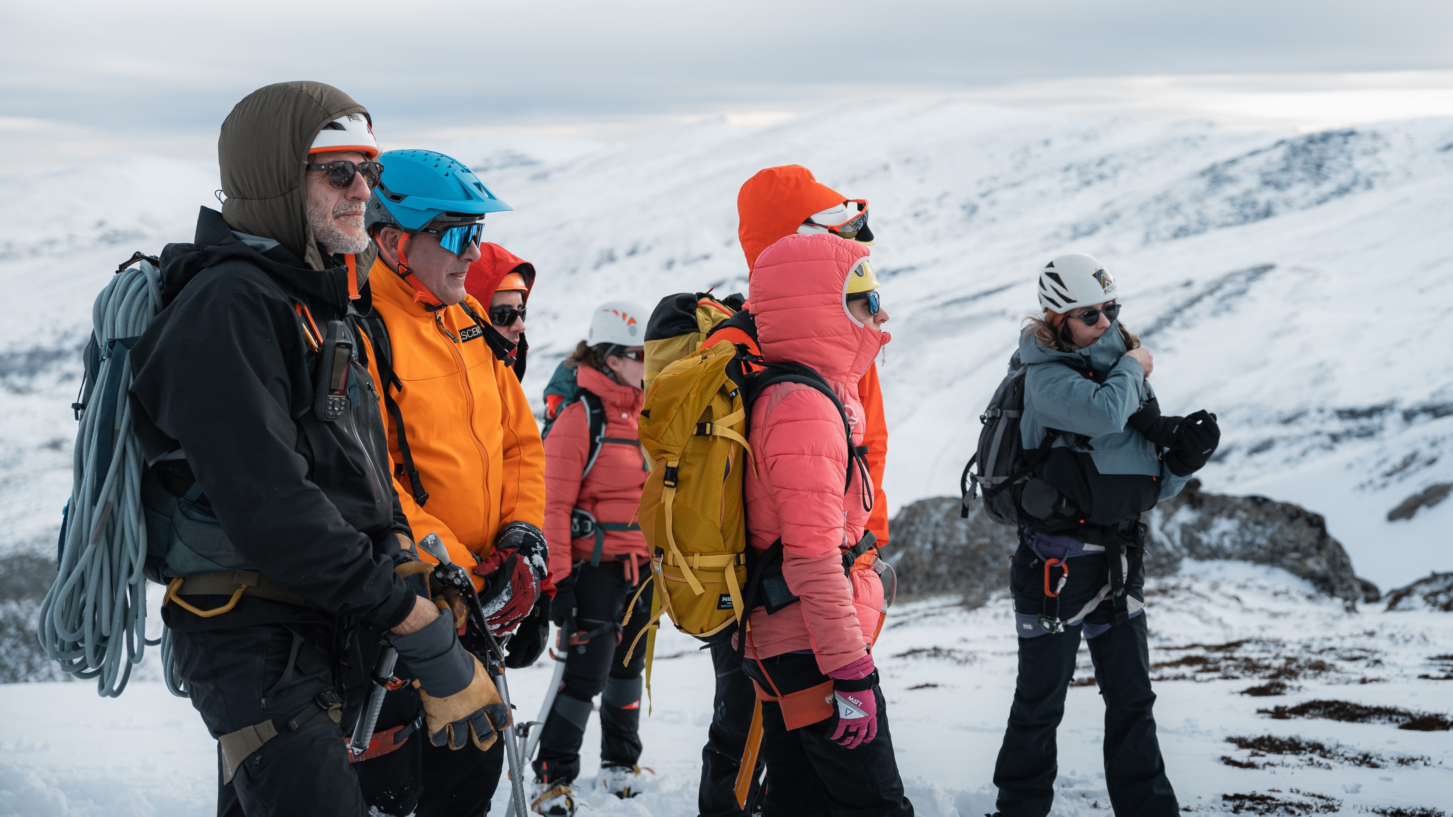 Grupo de esquiadores y alpinistas en un entorno nevado, con montañas al fondo y vistiéndose con equipos de invierno y protección adecuada.