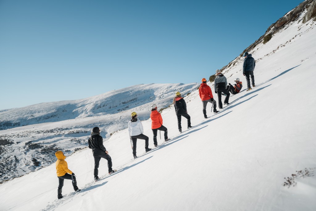 Un grupo de personas ascendiendo por una pendiente nevada en un día soleado, con las montañas cubiertas de nieve en el fondo.